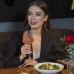 Home Elegant woman in a suit holding a cocktail, seated at a restaurant with food and decor.